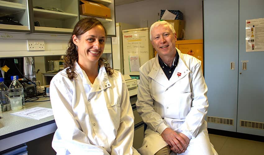 Two people wearing white laboratory coats smiling at the camera while seated on chairs in a science laboratory