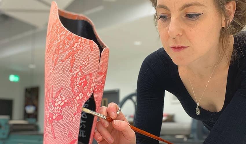 Woman working with a paintbrush on a pink coloured, patterned leg covering in a studio