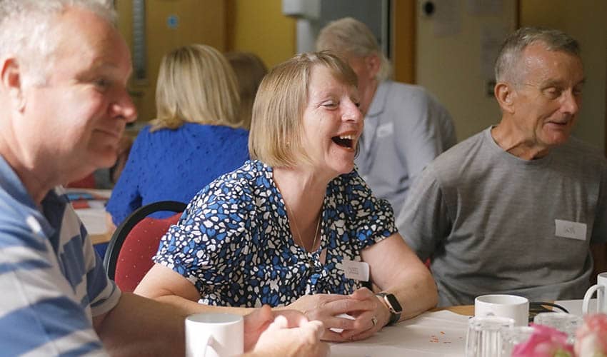 Photo of a dementia support celebration event, showing three people sat around a table. A lady in the middle of the photo is laughing.