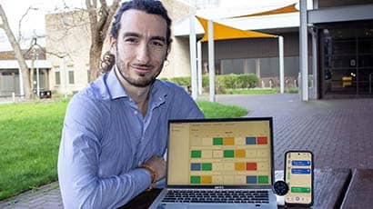 A man sat at a table outside a building, with a laptop computer, smartwatch and smartphone placed on the table displaying brightly coloured graphics
