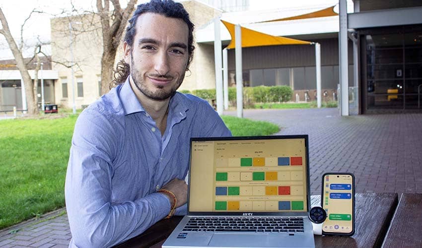 A man sat at a table outside a building, with a laptop computer, smartwatch and smartphone placed on the table displaying brightly coloured graphics