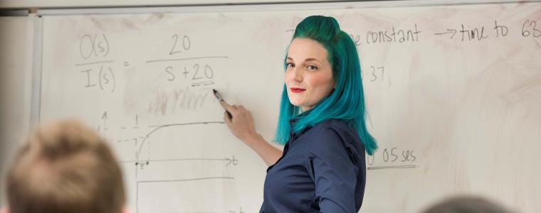 Woman with blue-green hair pointing to a white board displaying equations.