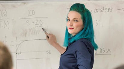 Woman with blue-green hair pointing to a white board displaying equations.