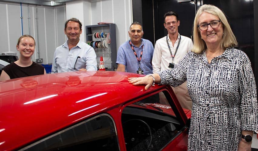 Five people standing and smiling at the camera, stood around a red Mini car