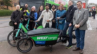 A  group of nine people stood behind a green cargo bike and a green electric bike, with a bus at a bus stop in the background