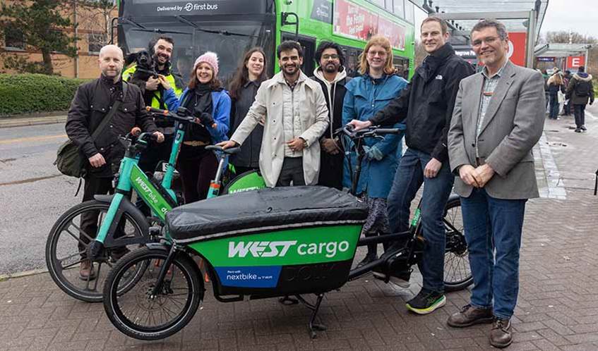 A group of nine people stood behind a green cargo bike and a green electric bike, with a bus at a bus stop in the background