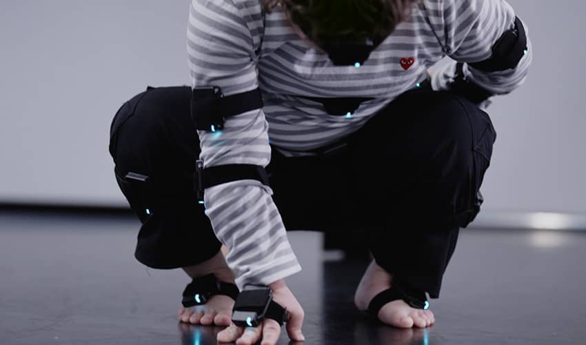 Annie Edwards crouching in a pose during a contemporary dance duet. Annie is wearing an inertial measurement motion capture suit, that is designed to capture her movements.
