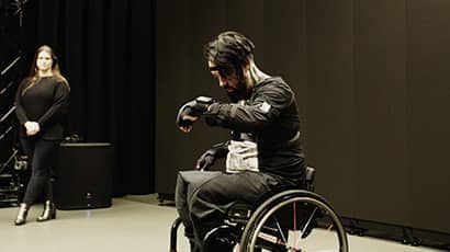Kris Halpin, a wheelchair user, performs music in a black box studio wearing a motion capture suit and data gloves, concentrating as he makes a gesture with his raised left hand.