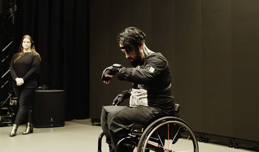 Kris Halpin, a wheelchair user, performs music in a black box studio wearing a motion capture suit and data gloves, concentrating as he makes a gesture with his raised left hand.