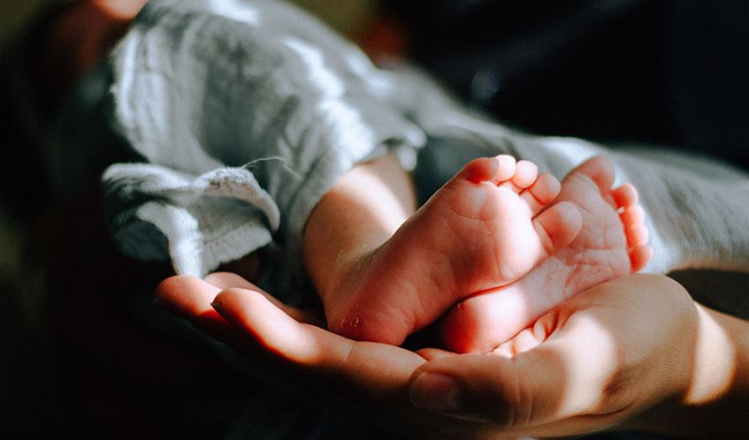Feet of a newborn baby, held by the hand of an adult, with a shard of sunlight shining onto both the feet and the hand