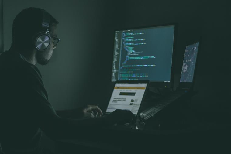 A person coding in a dark room using two laptops and a computer.