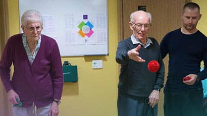 Older man and woman standing with physical trainer, man throwing a red ball.