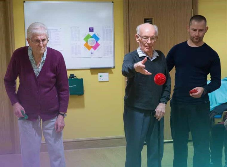 Older man and woman standing with physical trainer, man throwing a red ball.