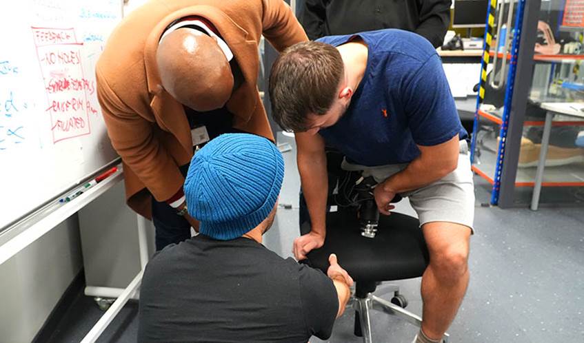 Image showing two male engineers fitting a prosthetic leg socket to the residual limb of a male amputee who is sat on a chair in a engineering laboratory at a university