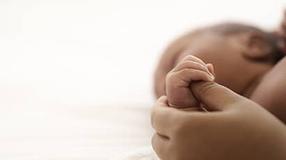Image of a newborn baby, with side of its face blurred in the background, and a parents hand holding the baby's hand in the foreground