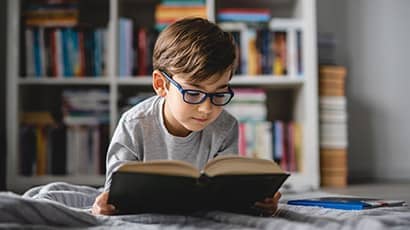 A boy wearing glasses reading a book while laying on a bed, with book shelves in the background