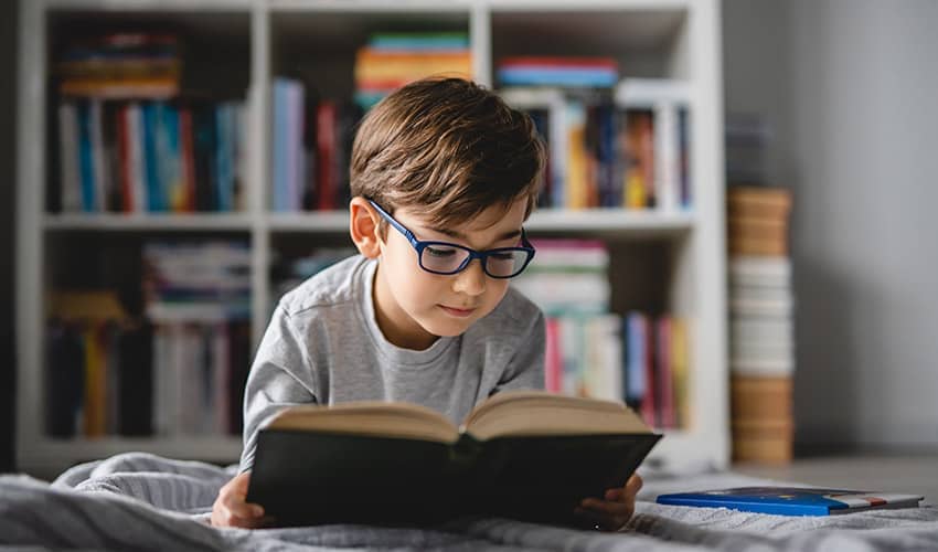 A boy wearing glasses reading a book while laying on a bed, with book shelves in the background