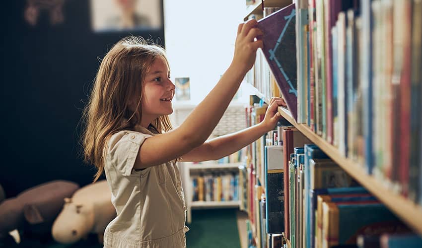 A girl smiling as she pulls a book from a book shelf in a library