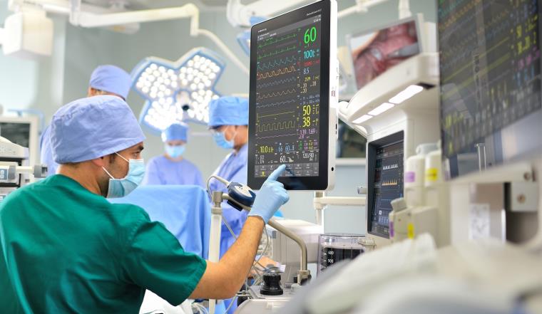 Man in green scrubs and face mask pressing large computer monitor.