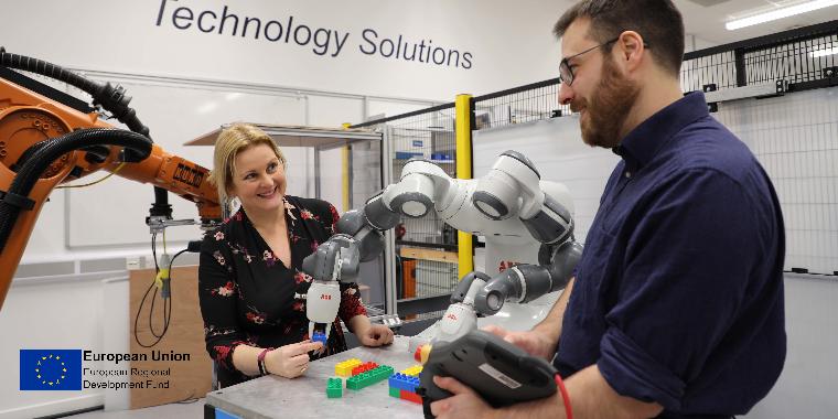 Man and woman stood either side of a large, white robotic arm.