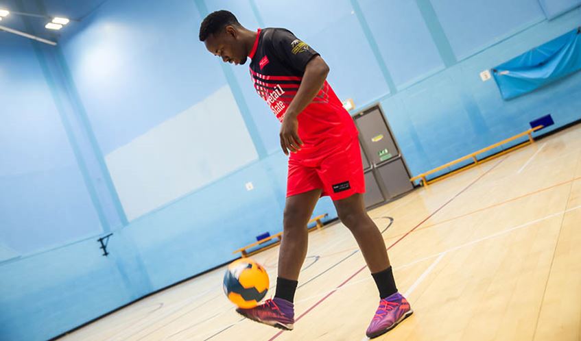 UWE Bristol international student Sadock John photographed in a sports hall wearing a red football kit kicking a yellow football