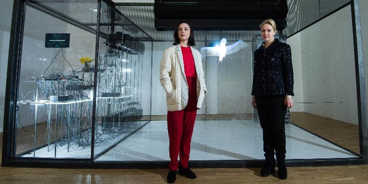 Two women standing in front of living bricks glass box gallery installation of many wires hanging from tables and screens.