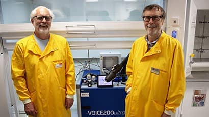 Two men in yellow lab coats smiling at the camera, standing in a science lab in front of a blue machine