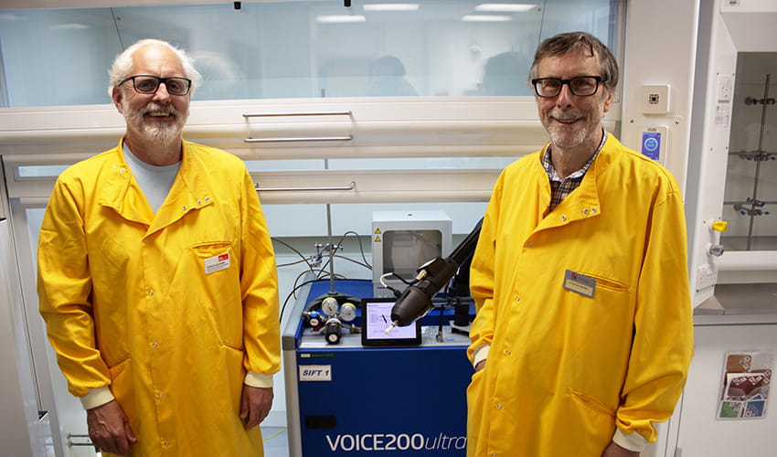 Two men in yellow lab coats smiling at the camera, standing in a science lab in front of a blue machine