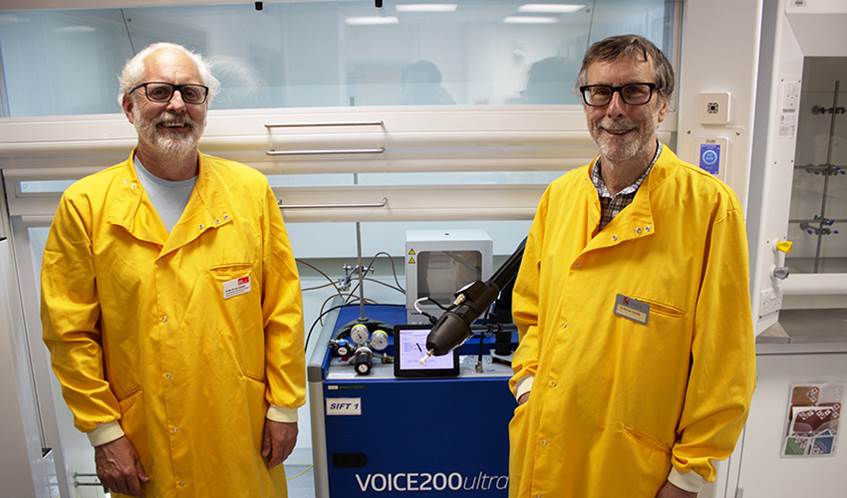 Two men in yellow lab coats smiling at the camera, standing in a science lab in front of a blue machine