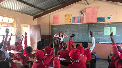 Teacher at front of classroom in front of blackboards and class of children in red uniforms holding hands up to answer a question. 