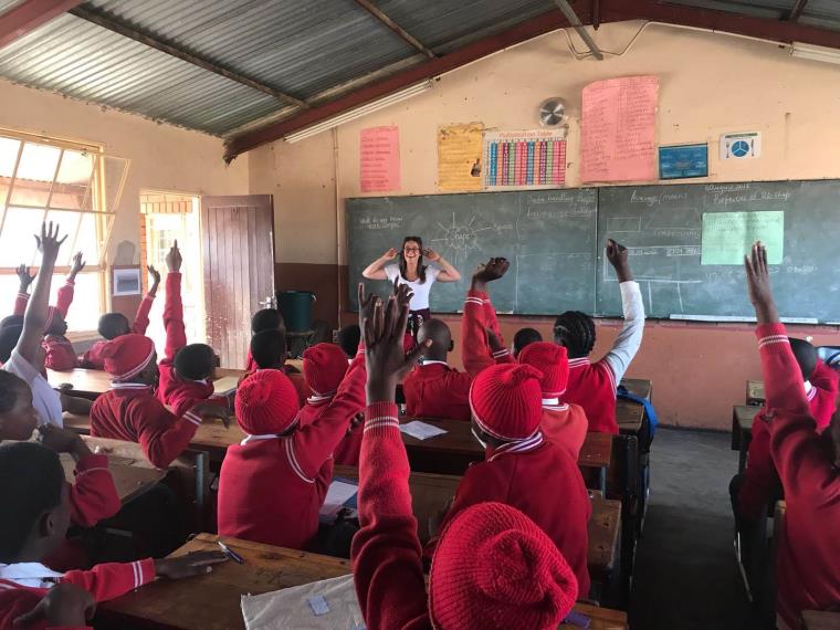Teacher at front of classroom in front of blackboards and class of children in red uniforms holding hands up to answer a question.