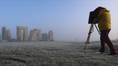 A photographer using an old-fashioned camera to take an photo of Stonehenge, with a backdrop of a blue sky