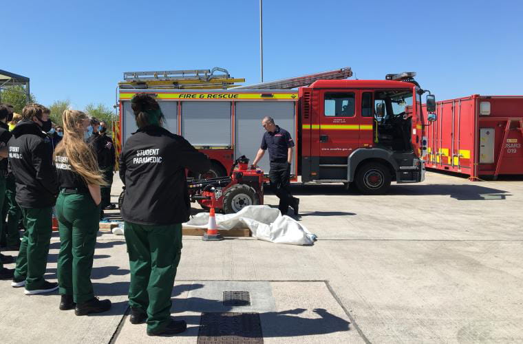 Student paramedics wearing green uniforms stand outdoors watching a demonstration of a red machine with wheels by a uniformed staff member from Avon Fire and Rescue standing in front of a fire engine.