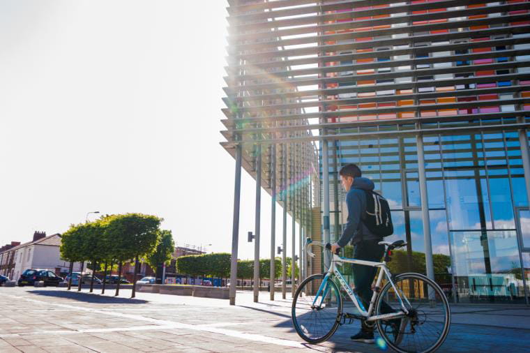 A student pushes a bicycle in front of a modern building.