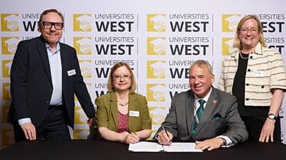 Four people at a desk smiling at the camera (two standing, two seated), with one of the people seated to the right signing a piece of paper in front of him