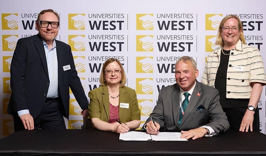 Four people at a desk smiling at the camera (two standing, two seated), with one of the people seated to the right signing a piece of paper in front of him