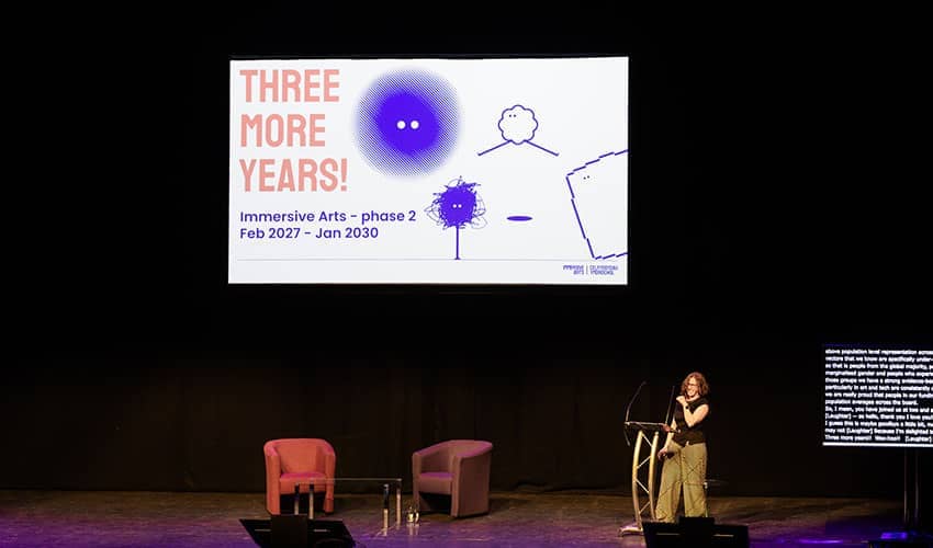 Verity McIntosh, Professor of Immersive Arts and Culture at UWE Bristol, speaking at an event beneath a large digital screen showing the text 'Three more years'