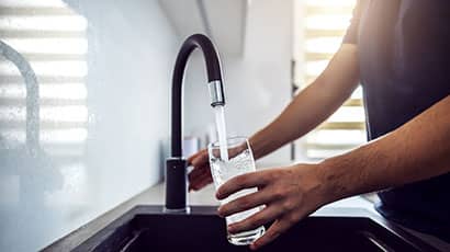 Image of a person filling up a glass with water from a tap, with the sun shining through a blind in the background