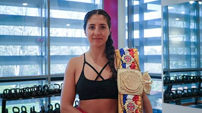A woman in a gym with a martial arts championship winner belt over her shoulder 