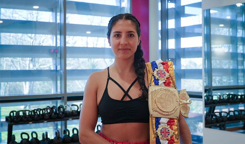 A woman in a gym with a martial arts championship winner belt over her shoulder