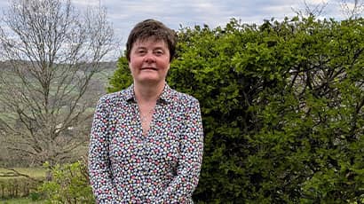 A woman stands smiling at the camera with open countryside behind her. 