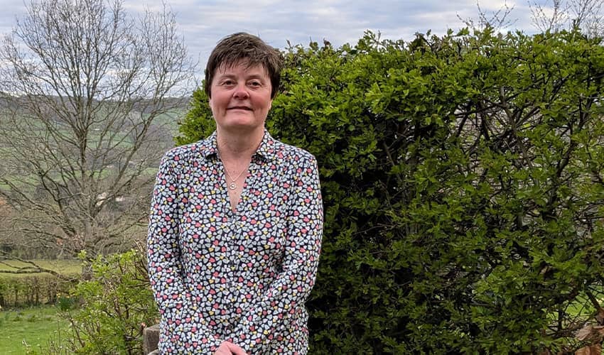 A woman stands smiling at the camera with open countryside behind her. 