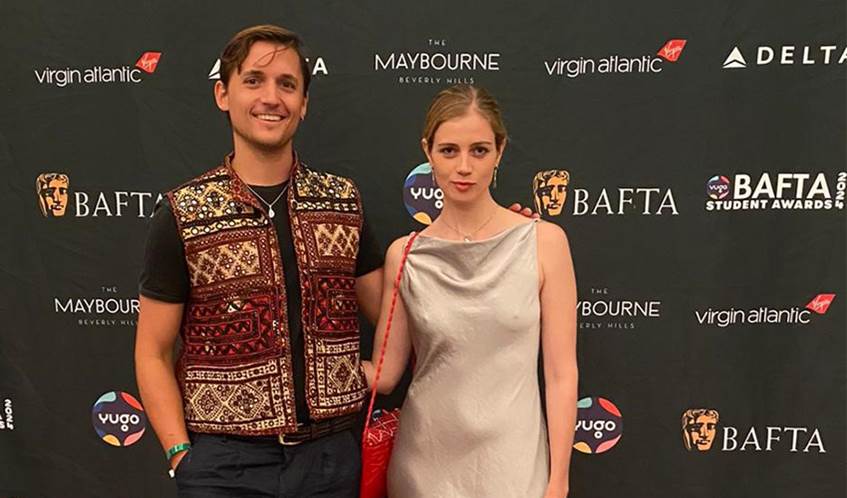 A man and woman stand smiling with an arm around each other looking at the camera.