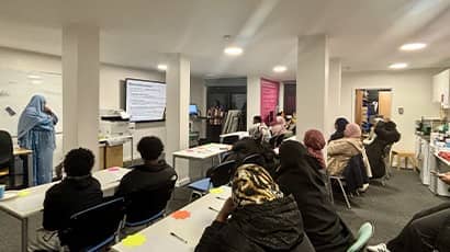 A classroom setting with two rows of tables with people sat at them facing a person stood at the front of talking to the group.