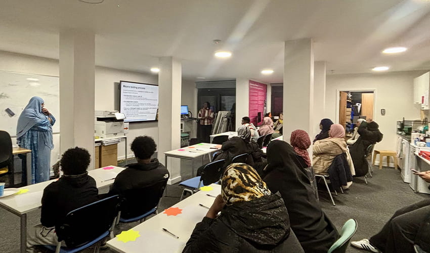 A classroom setting with two rows of tables with people sat at them facing a person stood at the front of talking to the group.