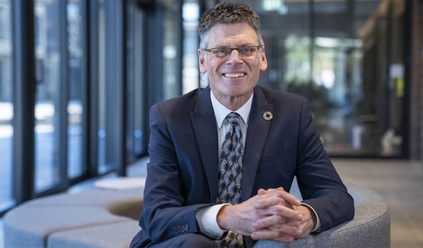 A headshot of a man in a blue suit in a corporate setting smiling at the camera.