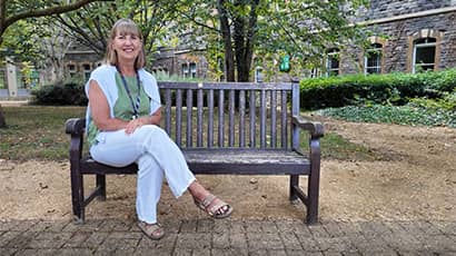 A woman sitting on a bench in front of trees, bushes and buildings. Windows can be seen in the background. The woman is wearing sandals, white trousers, a lanyard, a green top and has a white cardigan draped over her. 