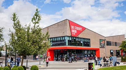 An exterior of a building with red branding with trees and a open paved walking space in front of it. 