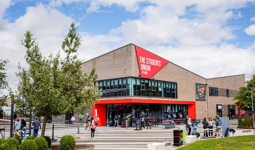 An exterior of a building with red branding with trees and a open paved walking space in front of it.