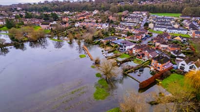 A birds eye view image taken from height overlooking houses which have been flooded. In front of the houses is a large field which is flooded with water. 